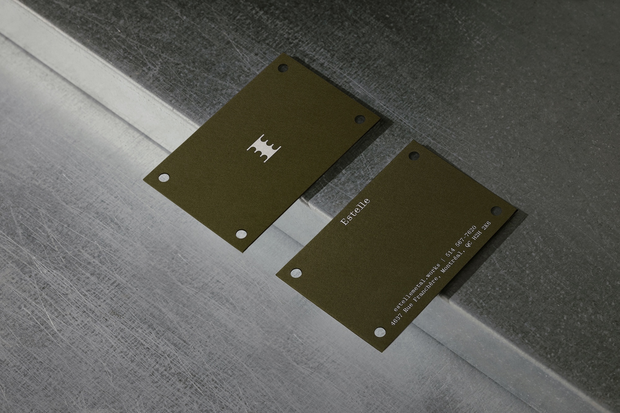 Two olive green metal cards with punched corner holes resting on a brushed steel and concrete surface.