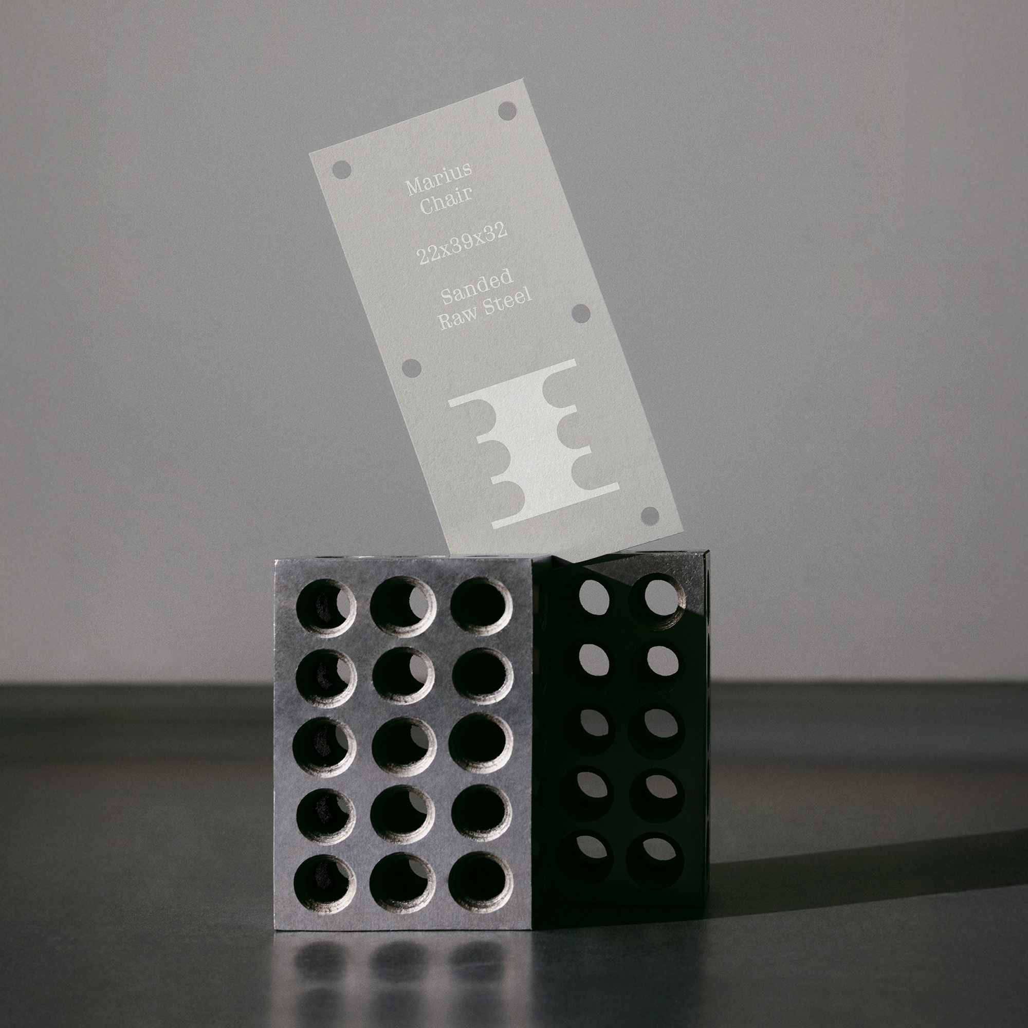 Perforated raw steel blocks with a semi-transparent branded product tag resting against them on a grey studio background.