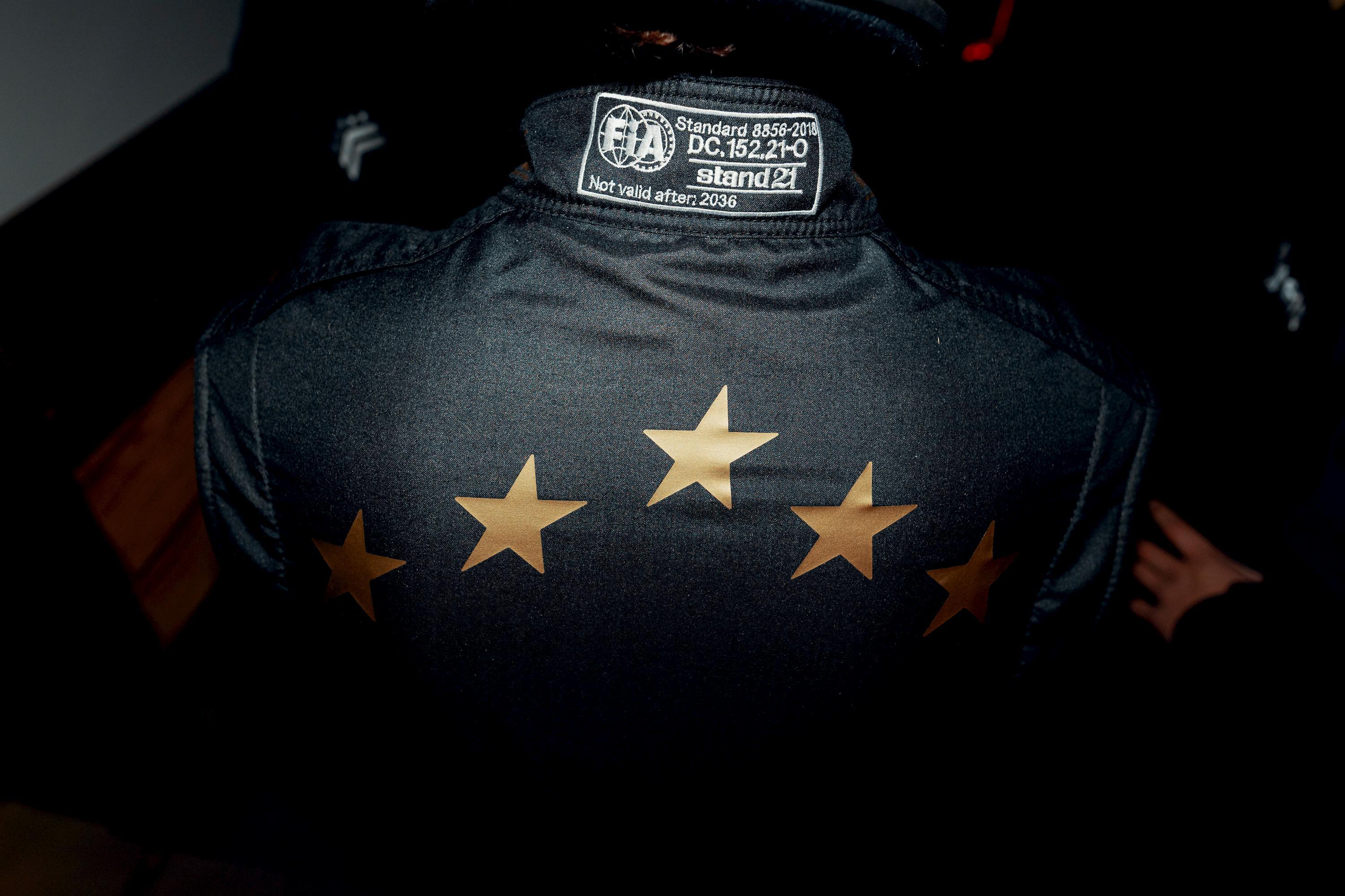 Rear view of a person wearing a black racing suit with a row of five gold stars and an FIA homologation collar tag.
