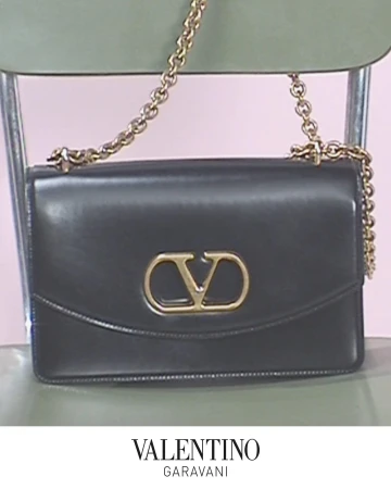 Black leather shoulder bag with gold chain strap and metal V logo resting on a green chair