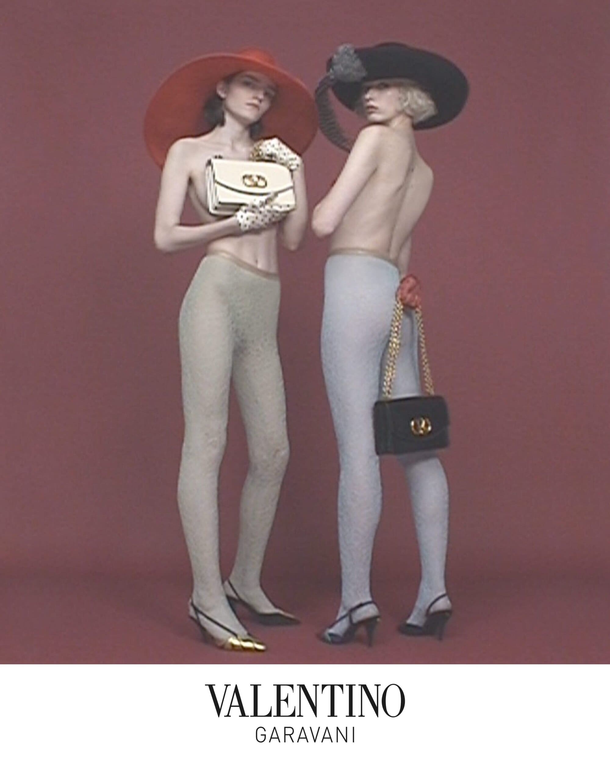 Two fashion models wearing wide-brimmed hats and designer handbags against a muted burgundy studio background for Valentino.