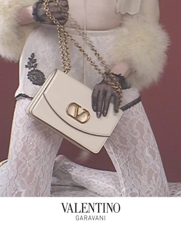 Model holding a white leather Valentino Garavani Vain bag with a gold chain strap against a red wall.