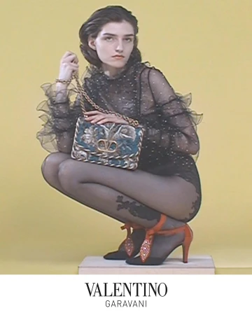 Model in black ruffled top holding an embroidered Valentino Garavani chain bag against a yellow studio backdrop.