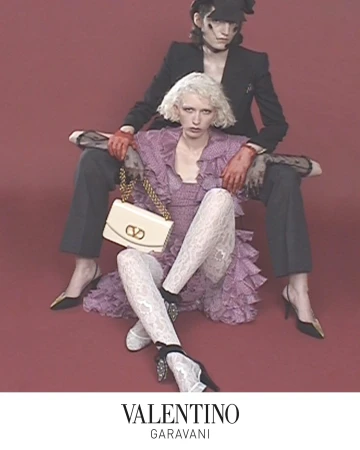 Two models posing against a solid red backdrop, featuring a white Vain handbag and vintage-styled attire.