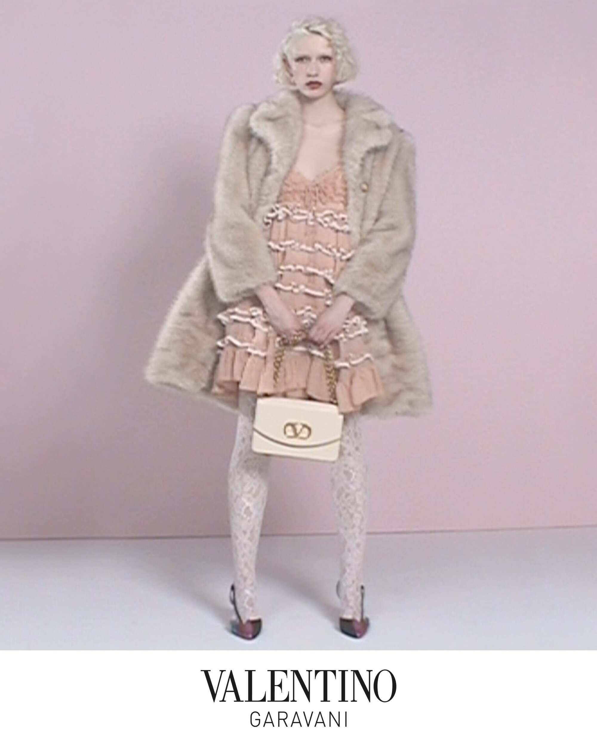 Model in ruffled dress and faux fur coat holding a white handbag against a pale background