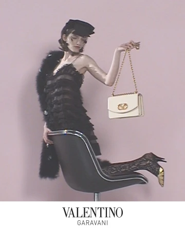 Model in black ruffle dress holding a white handbag while leaning over a chair against a pink backdrop