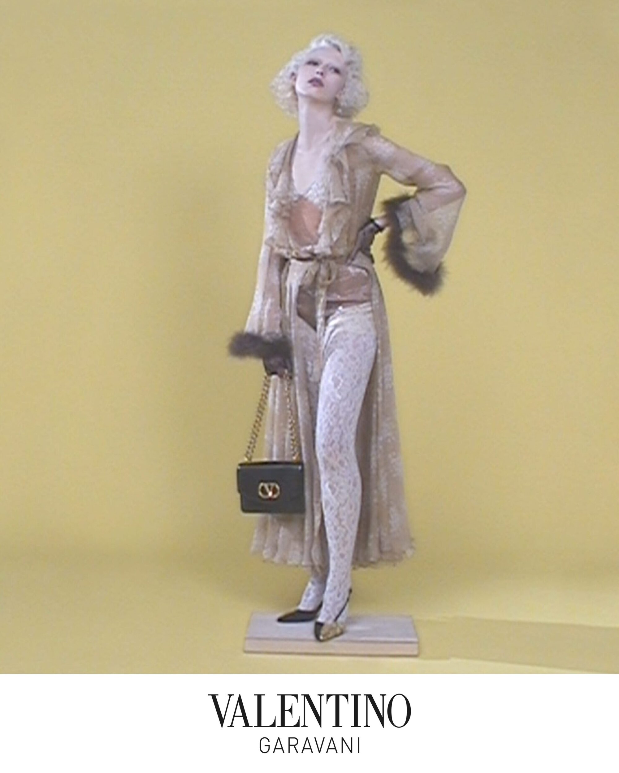 Model with blonde hair wearing sheer ruffled robe holding a black chain strap handbag against a solid yellow backdrop