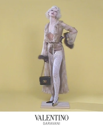 Model with blonde hair wearing sheer ruffled robe holding a black chain strap handbag against a solid yellow backdrop