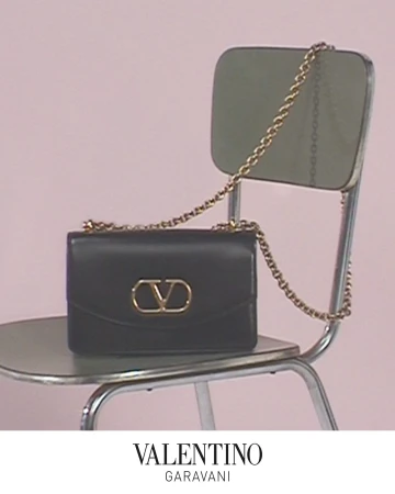 Black leather shoulder bag with a gold chain strap resting on a metal chair against a pink background