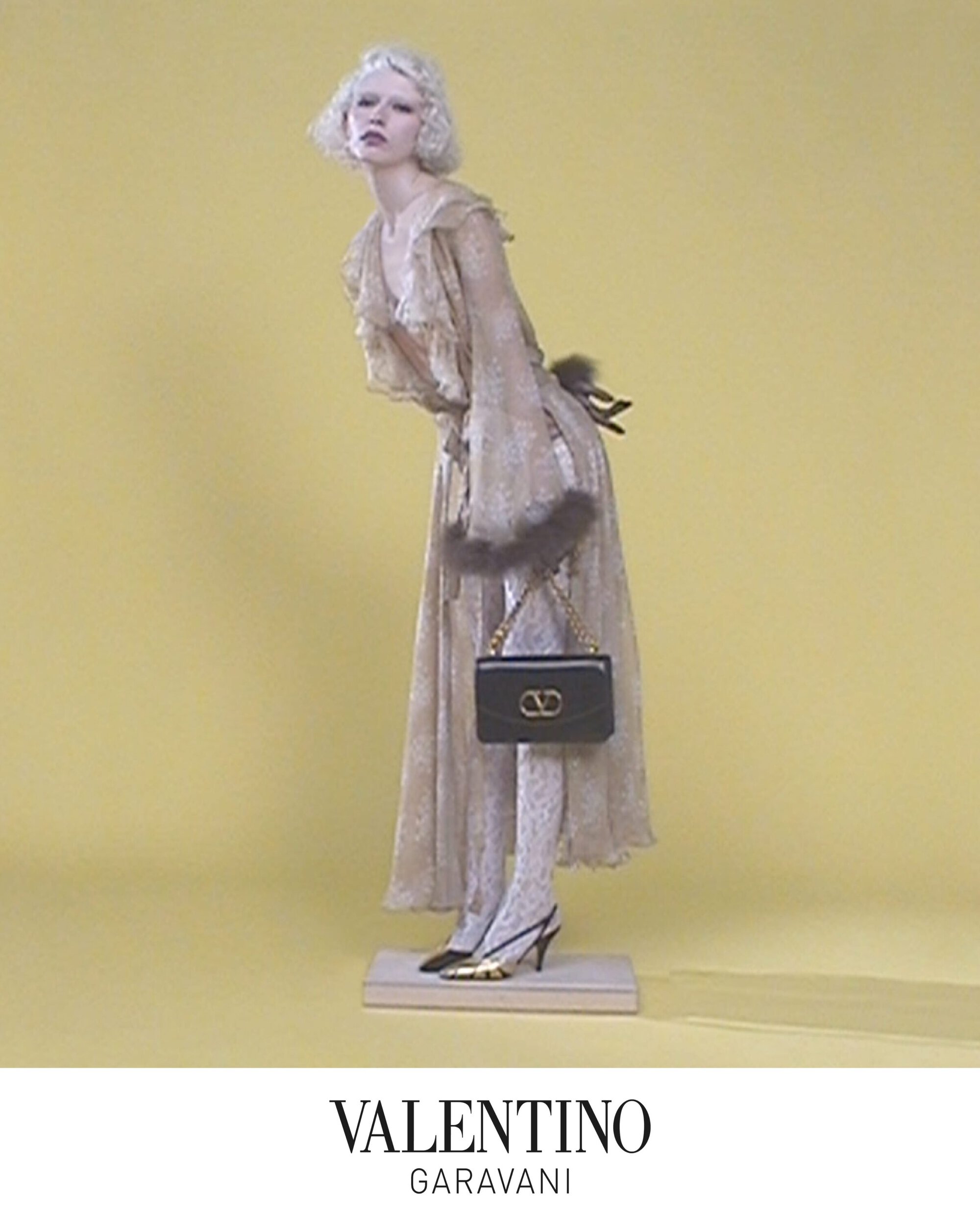 A blonde model wearing patterned dress and holding a black handbag against a solid yellow backdrop for Valentino