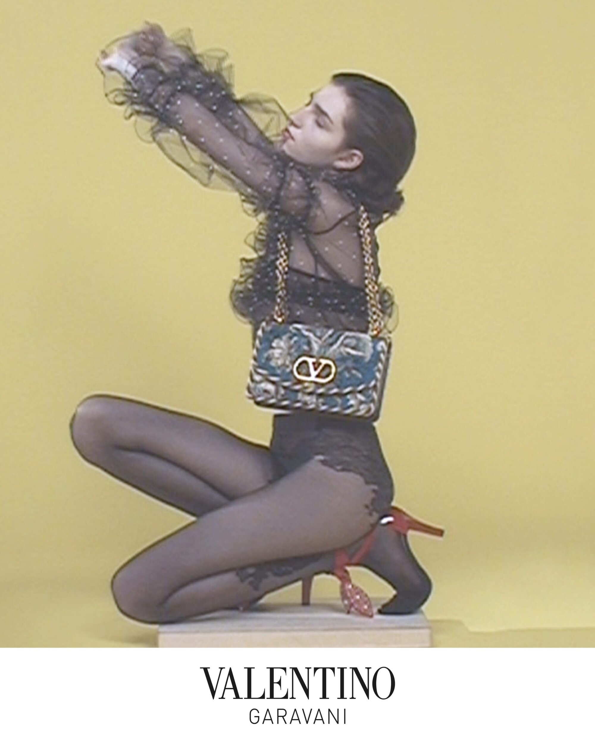 Model kneeling in black sheer top and patterned shoulder bag against a solid yellow background