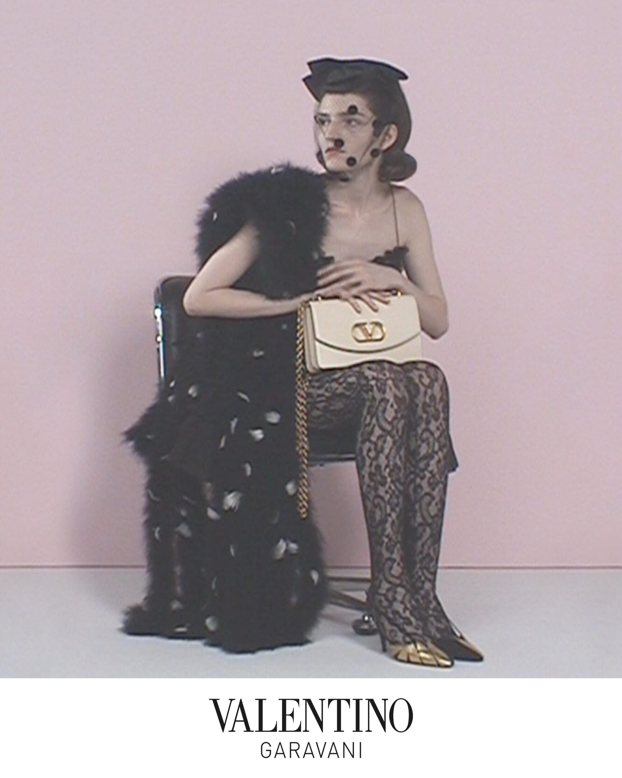 Model seated on a chair wearing a black feathered coat and patterned tights while holding a cream handbag against a pink background