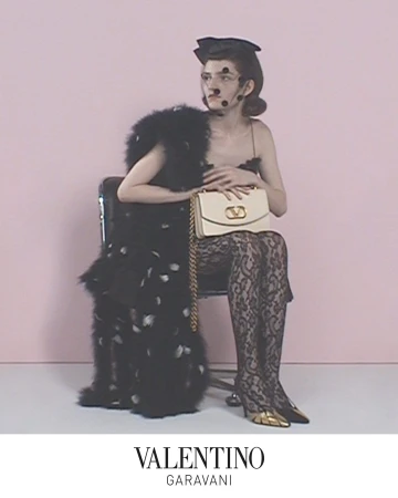 Model seated on a chair wearing a black feathered coat and patterned tights while holding a cream handbag against a pink background