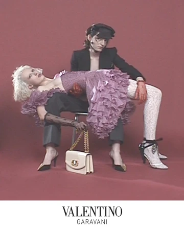 Two models posed against a muted red backdrop with a cream handbag resting on the floor in front of them.