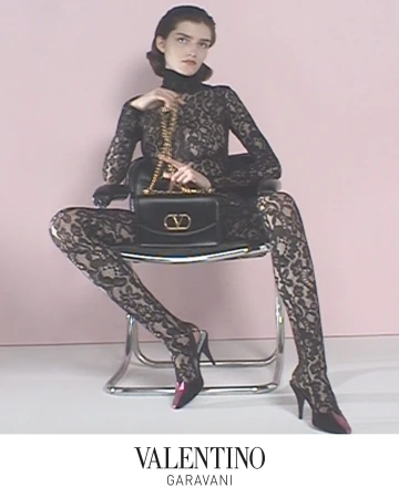 Model in black lace catsuit seated on a chair holding a black handbag with gold hardware against a light background