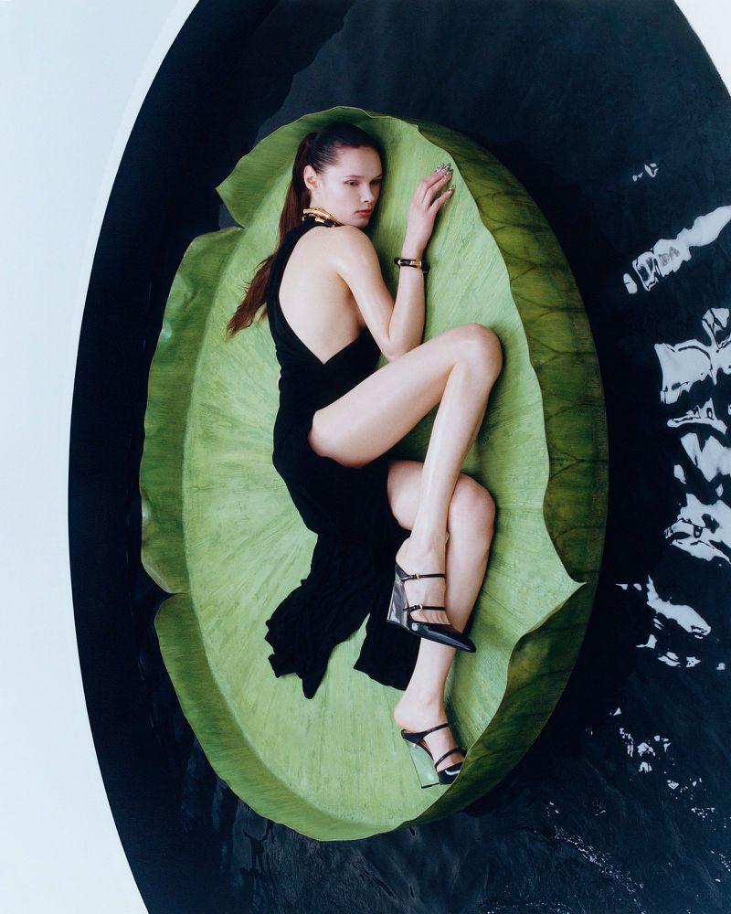 Woman in black dress lounges on a large green leaf surrounded by water in a fashion editorial, showcasing beauty and elegance.