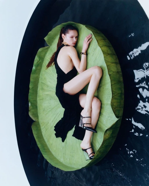 Woman in black dress lounges on a large green leaf surrounded by water in a fashion editorial, showcasing beauty and elegance.