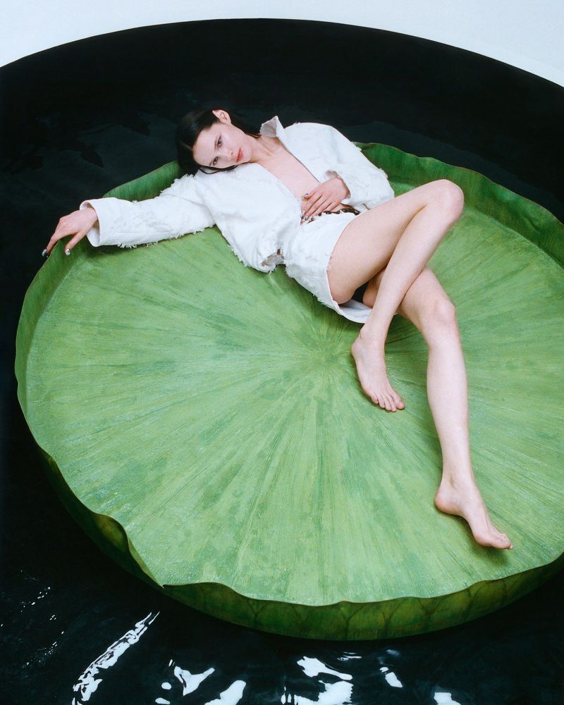 Woman in a white jacket and shorts lies on a large green lily pad surrounded by black water in a studio setting, looking at camera.