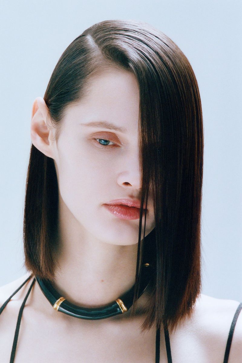 Brunette woman with striking dark bob haircut and light blue eyes posing against a light backdrop, wearing a thick black necklace.