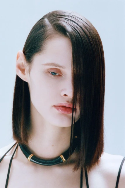 Brunette woman with striking dark bob haircut and light blue eyes posing against a light backdrop, wearing a thick black necklace.