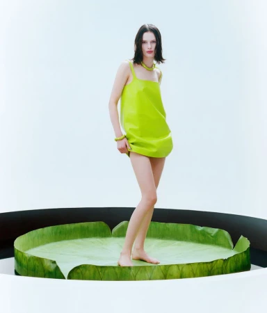 Fashion shot of a young adult female model with bare feet, wearing a green dress, standing on a giant green leaf in a white environment.