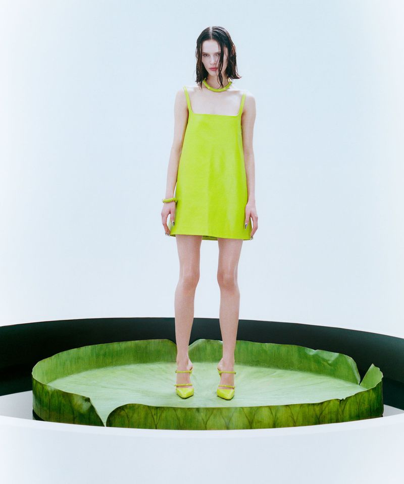 Fashion model poses in a vibrant yellow mini dress, necklace, bracelet, and shoes, standing on a large green leaf-shaped platform.
