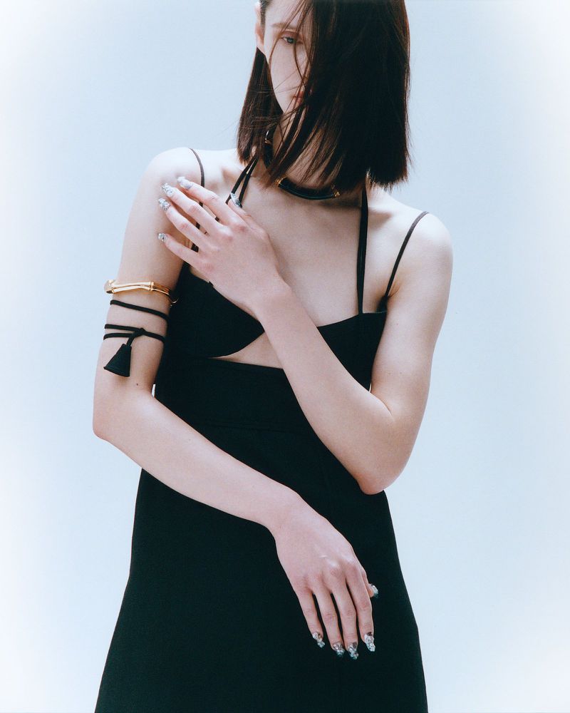 A fashionable adult female model poses indoors wearing a black dress with minimalist jewelry against a bright, neutral background.