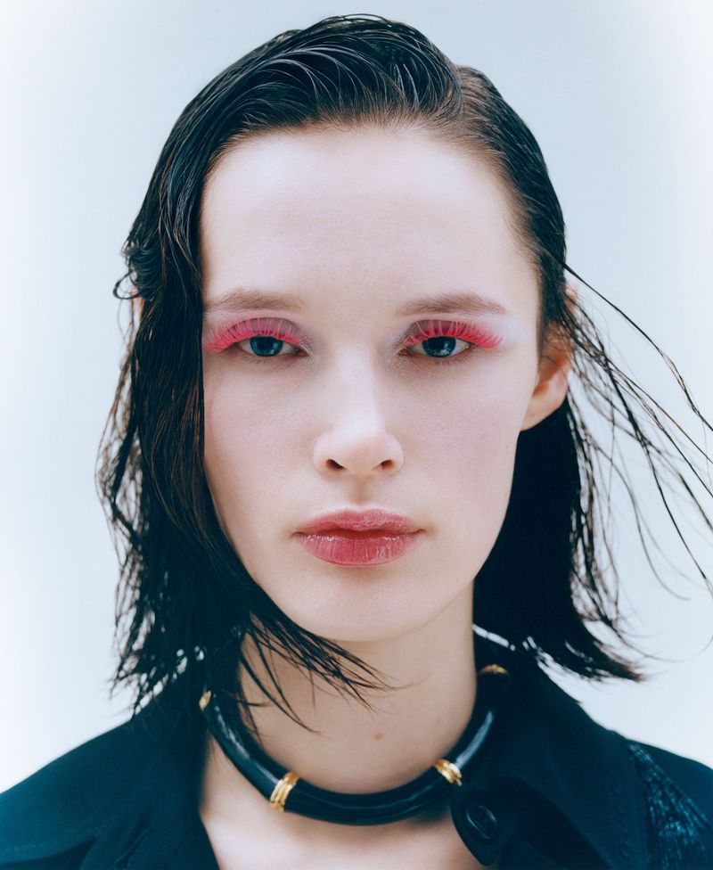 Headshot of a woman with wet, dark hair, pink makeup on her eyelashes and lips, wearing a black choker and a black jacket against a white background.