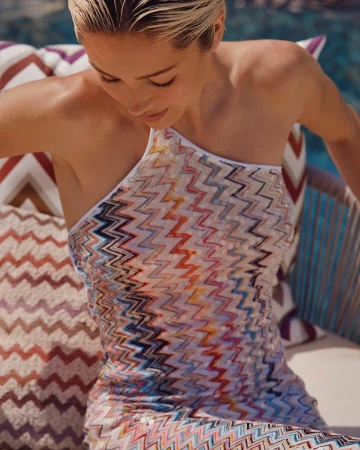 Model wearing a multi-colored zigzag patterned one-shoulder dress outdoors near a pool with patterned cushions.