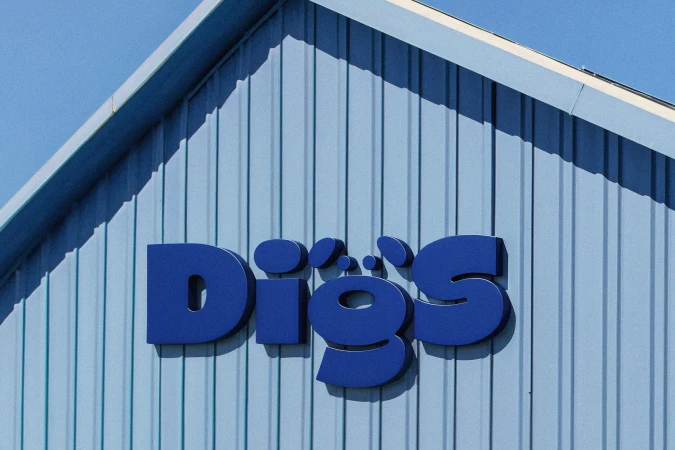Blue metallic letters spelling DIGS mounted on a light blue vertical corrugated metal building exterior.