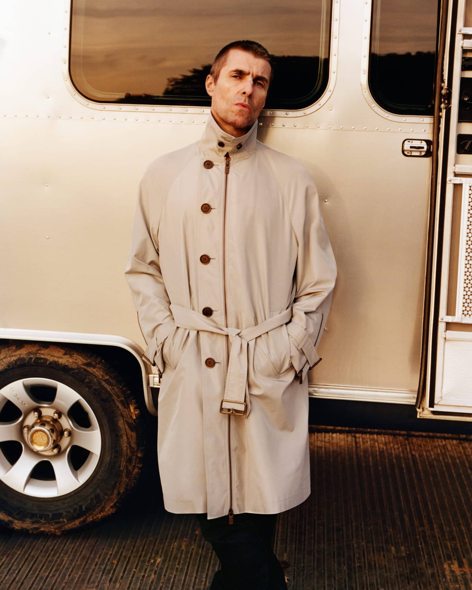 Liam Gallagher wearing a light-coloured belted trench coat leaning against a metallic trailer for Burberry