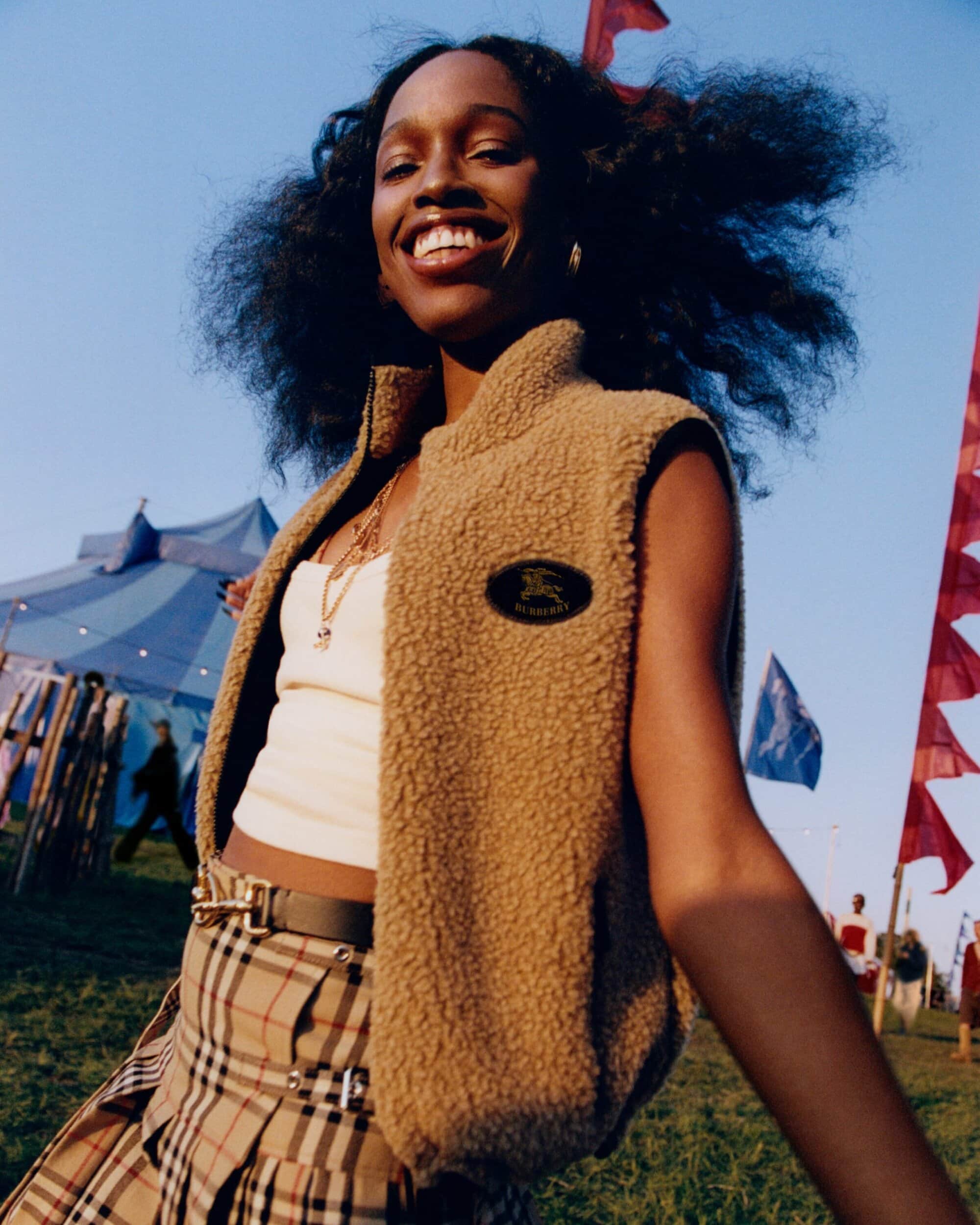 Smiling woman with voluminous dark hair wearing a camel fleece vest and plaid skirt at an outdoor festival with tents and flags
