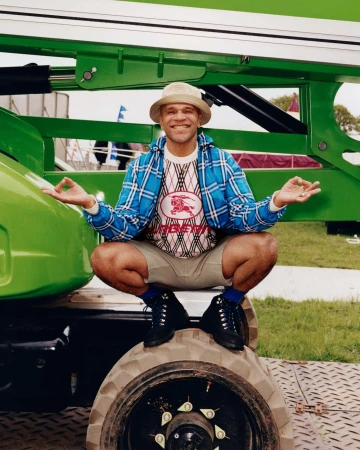 Goldie squatting on a heavy machinery tire wearing a blue plaid jacket and bucket hat for Burberry Festival Ad Campaign.