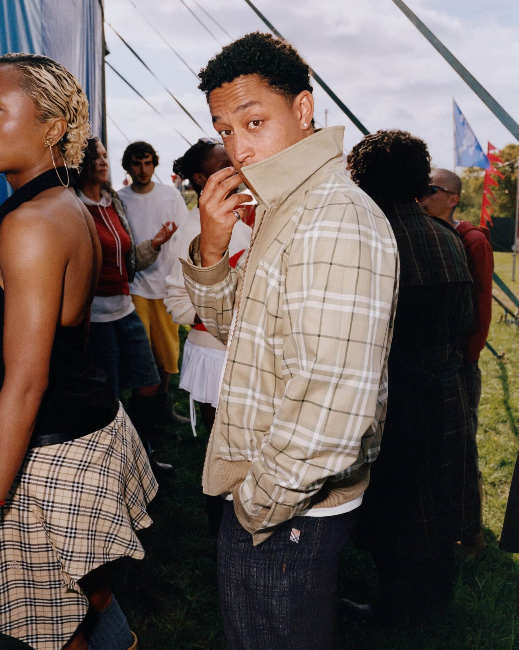 A man wearing a tan plaid check jacket with his collar popped stands outdoors near a tent structure with a group of people.