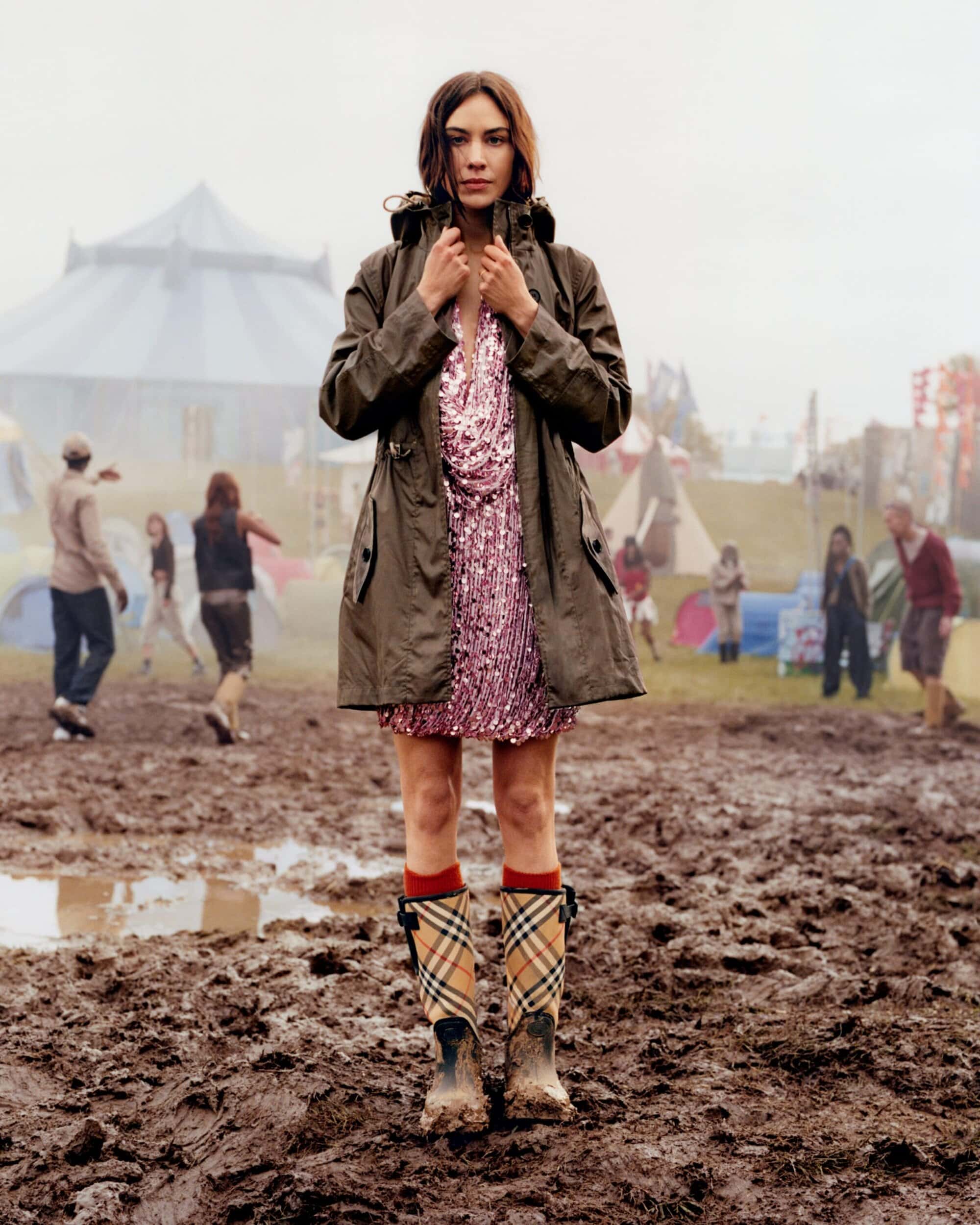Alexa Chung wearing an olive green jacket and patterned wellington boots standing on muddy ground at a festival