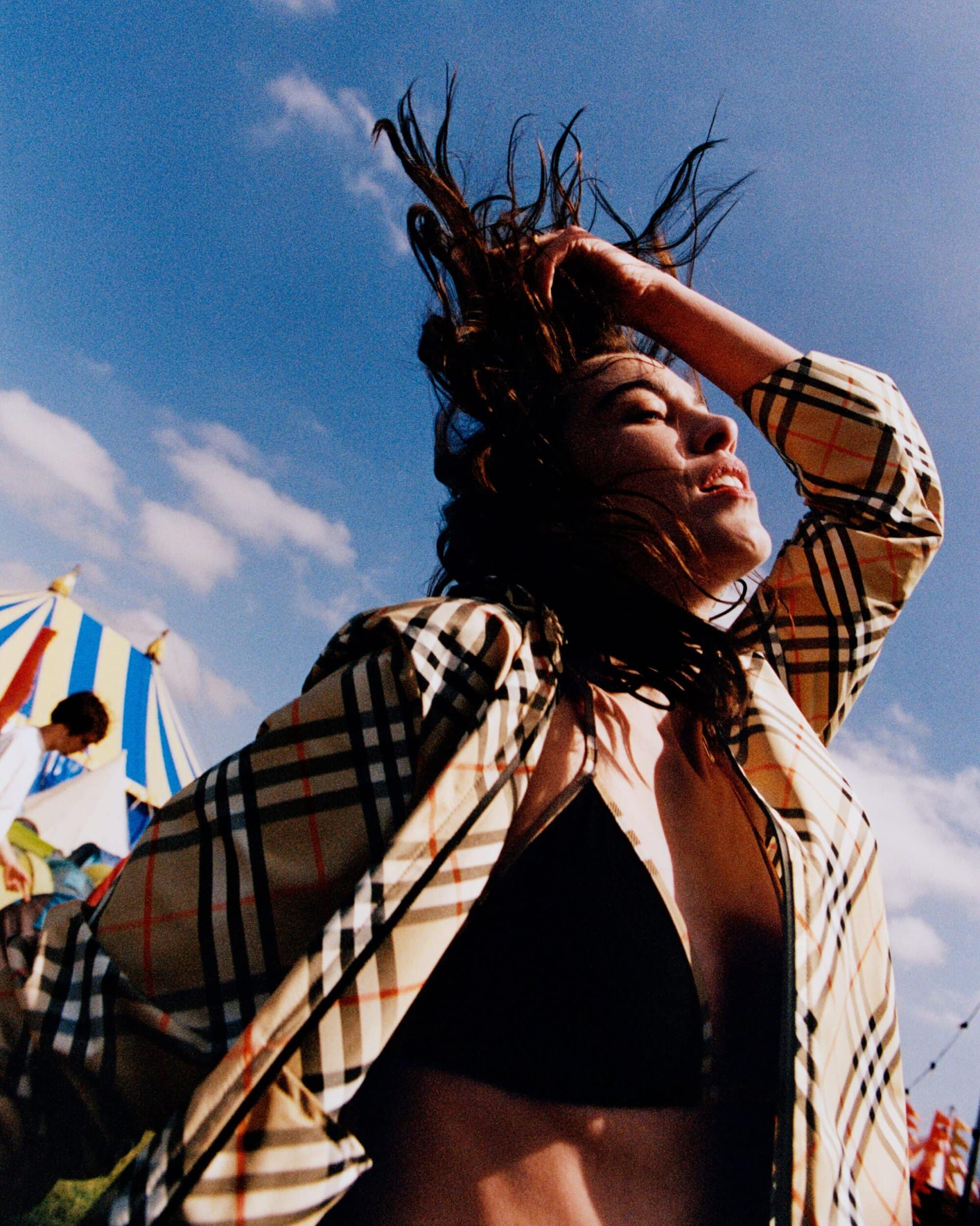 Low-angle shot of a woman with dark hair tossing her head back, wearing a Burberry check jacket outdoors under a blue sky.
