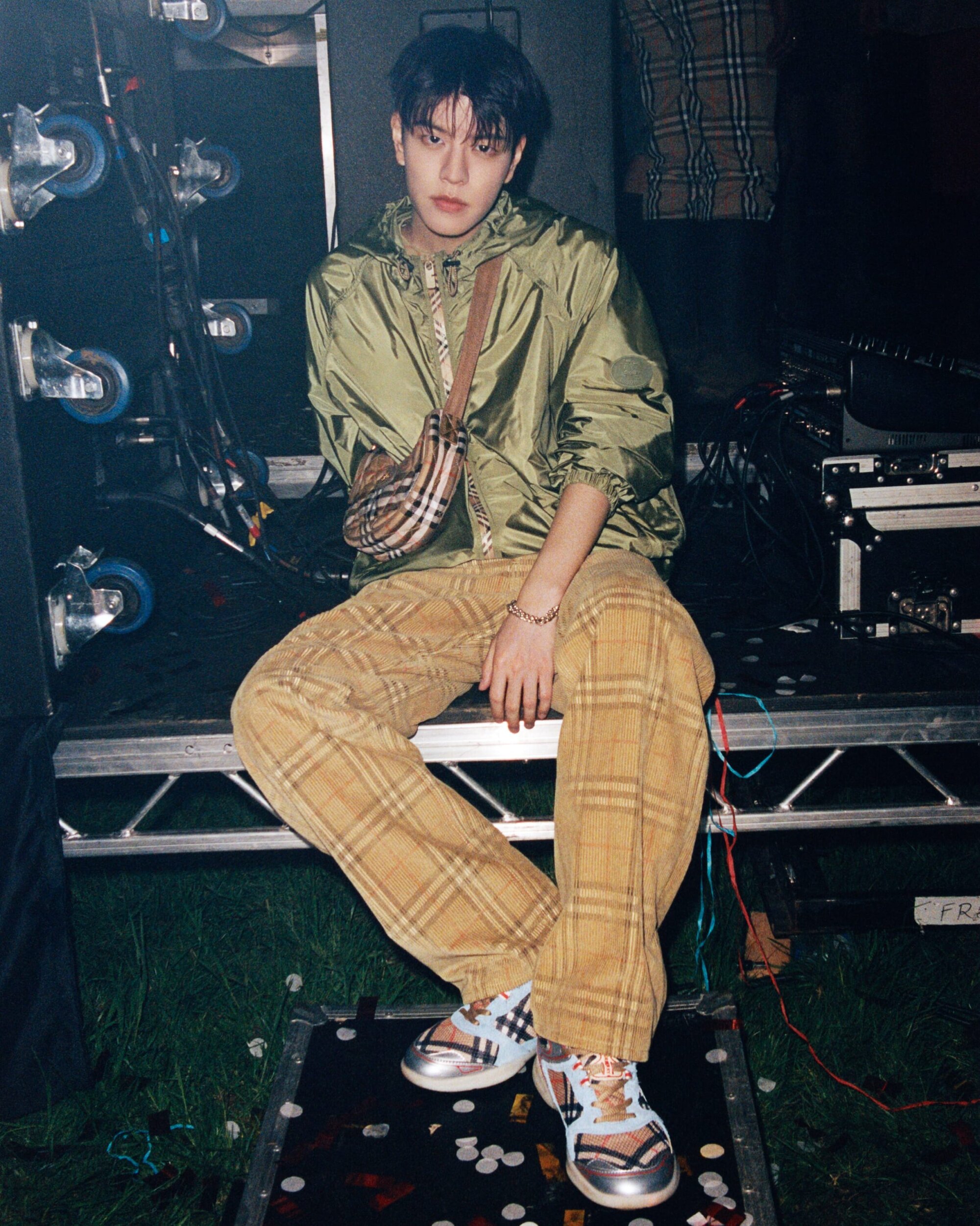 SEUNGMIN wearing a green jacket and plaid trousers seated on backstage stage equipment for Burberry festival ad campaign