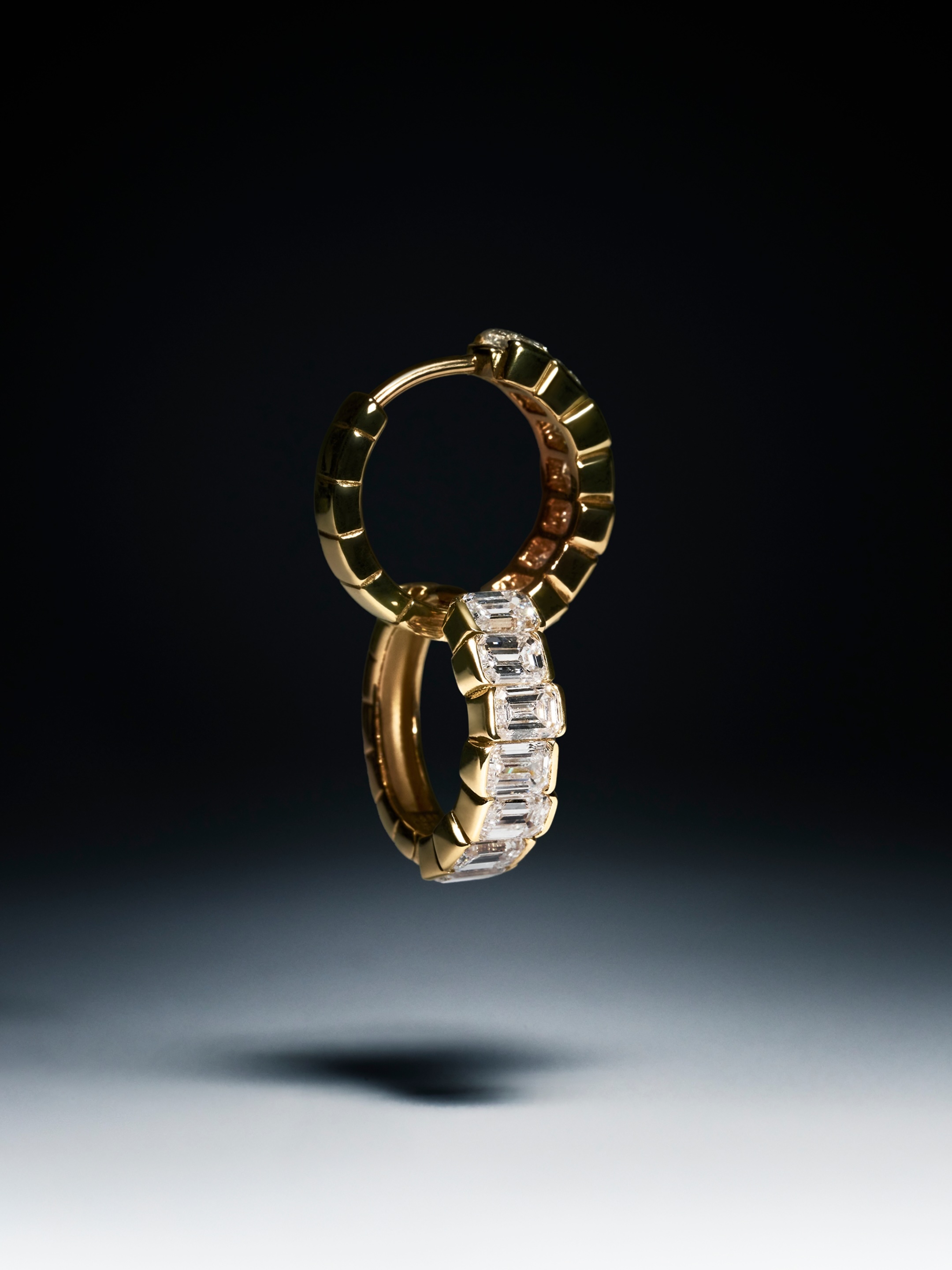 Two interlocking gold hoop earrings set with emerald-cut diamonds floating against a dark studio gradient background.