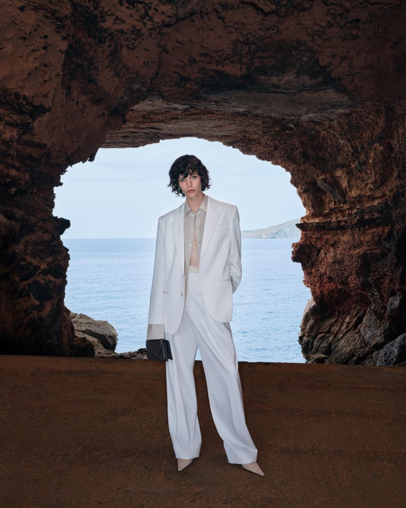 Model standing inside a rocky cave opening wearing a white suit against an ocean and sky backdrop for Victoria Beckham High Summer campaign