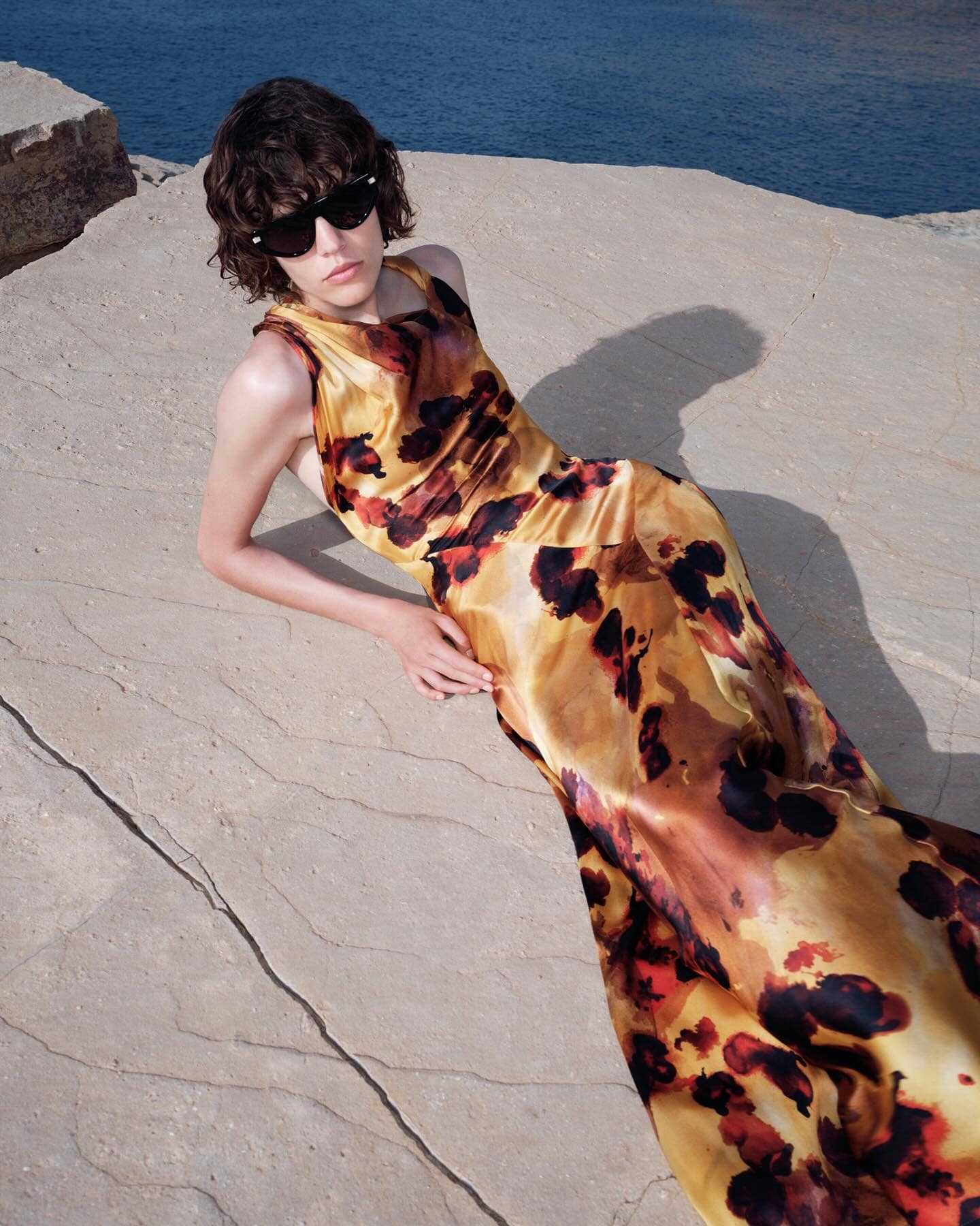 Model in printed yellow dress and black sunglasses reclining on rocky coastal stone surface near blue water