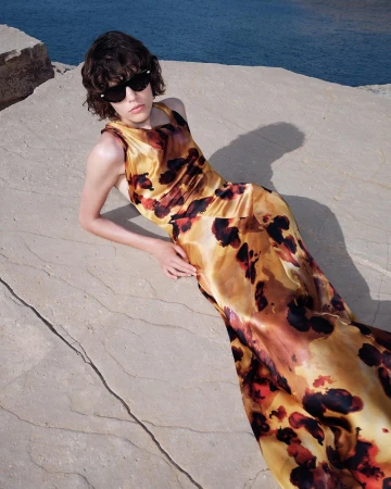 Model in printed yellow dress and black sunglasses reclining on rocky coastal stone surface near blue water