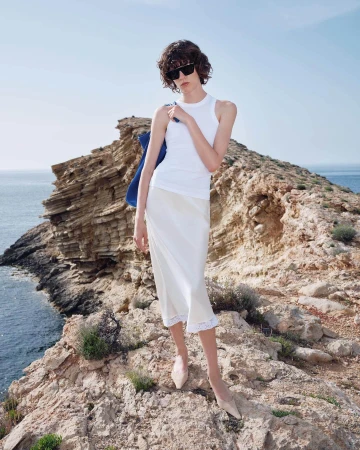 Model in white top and light skirt standing on rocky coastal cliff above the ocean wearing sunglasses.