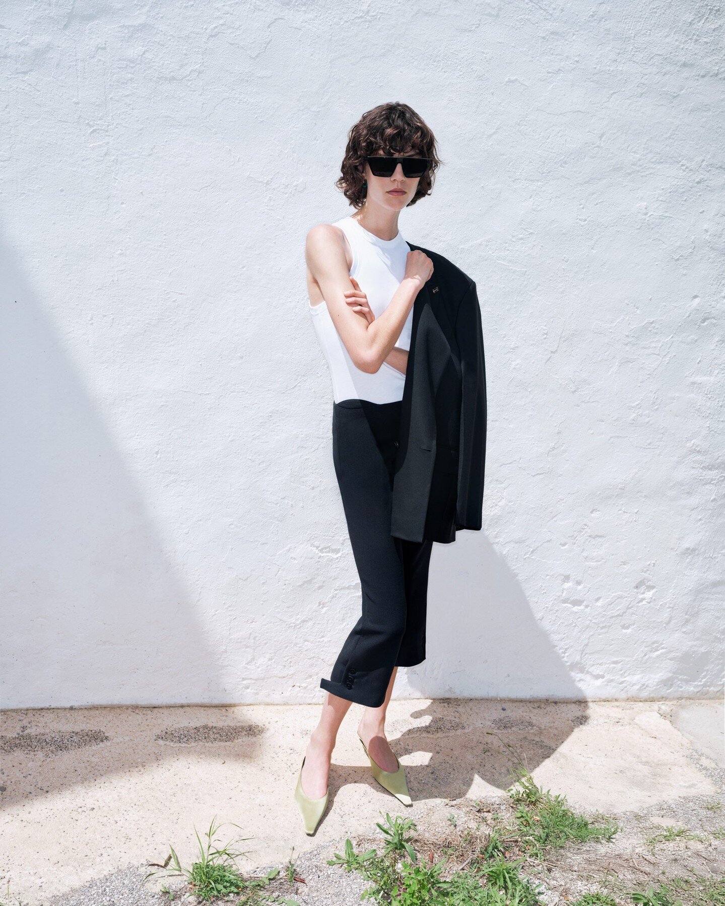 Model in dark sunglasses holding a black jacket against a white textured wall