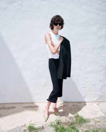 Model in dark sunglasses holding a black jacket against a white textured wall