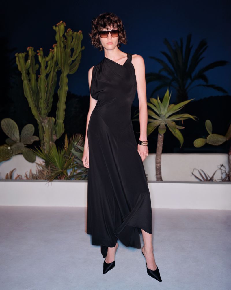 Model wearing a black sleeveless dress and sunglasses standing outdoors at night with cacti and palm trees.