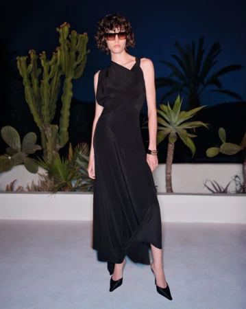 Model wearing a black sleeveless dress and sunglasses standing outdoors at night with cacti and palm trees.