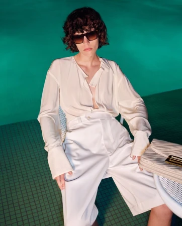 Model wearing dark sunglasses and cream silk blouse by Victoria Beckham against a turquoise pool background