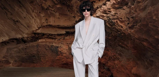 Model wearing a light grey double-breasted suit and black sunglasses posing against a rocky cavern wall.