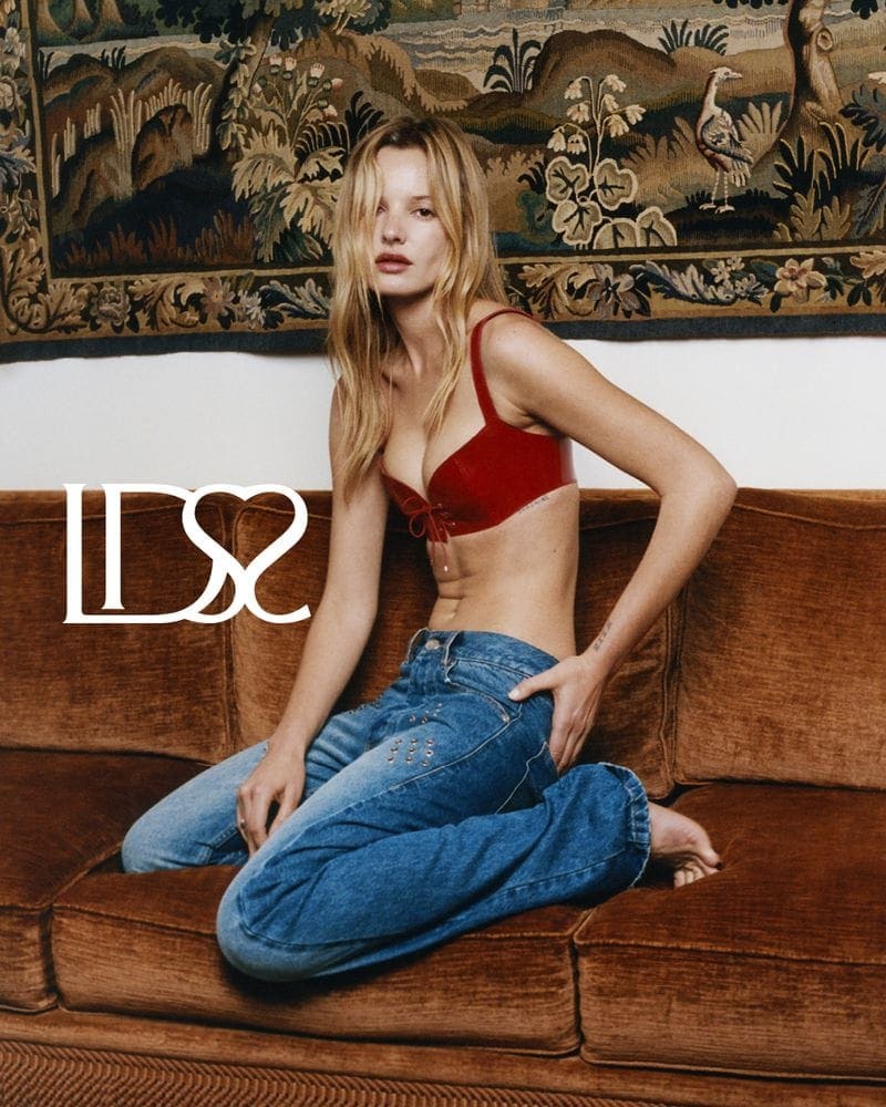 Model kneeling on a brown velvet sofa wearing a red bra top and blue jeans against a tapestry background for Ludovic de Saint Sernin.