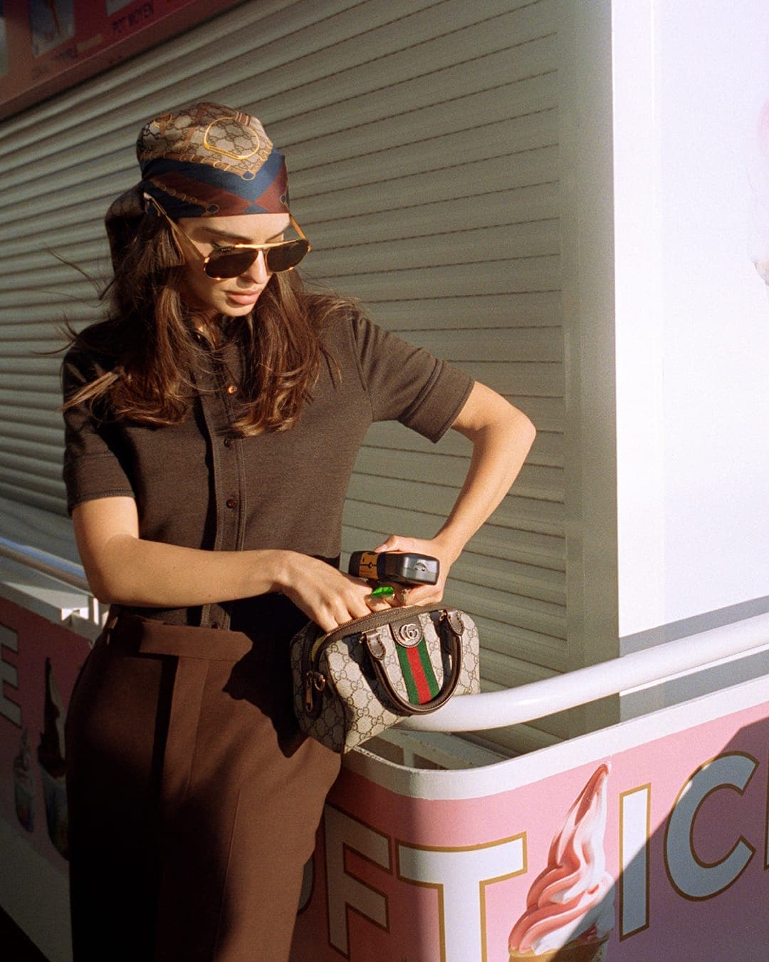 Model wearing a monogram headscarf and sunglasses holding a Gucci monogram handbag against a shuttered kiosk